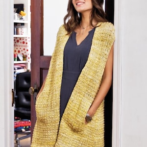 May include: A long, sleeveless, yellow knitted vest with pockets. The vest has a subtle speckled pattern and is worn over a dark grey dress.