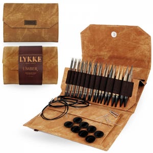 May include: A brown leather case with a zipper closure, containing a set of interchangeable knitting needles. The case has the text "LYKKE MAKE HAPPY UMBER" printed on the front. The needles are arranged in rows and are made of metal with black tips. The case also includes a set of black buttons, a pair of scissors, and a few lengths of black yarn.