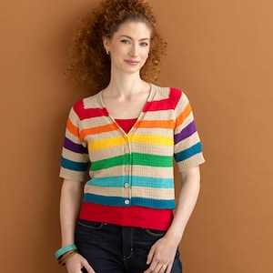 Op de afbeelding: Een gestreepte cardigan met korte mouwen in regenboogkleuren: rood, oranje, geel, groen, blauw en paars. De cardigan heeft een knoopsluiting en wordt gedragen met een rode top en een donkerblauwe jeans. De achtergrond is bruin.