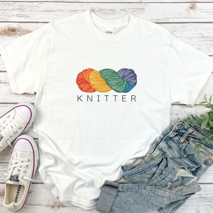 Puede incluir: Camiseta blanca con un gr&aacute;fico de ovillo de lana arco&iacute;ris y el texto "KNITTER" impreso en la parte delantera.