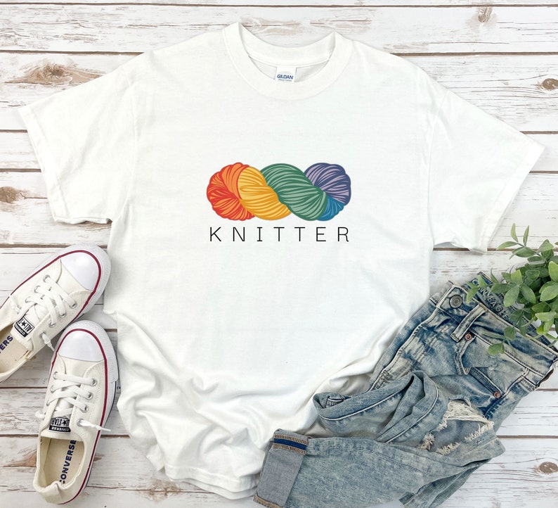 Puede incluir: Camiseta blanca con un gr&aacute;fico de ovillo de lana arco&iacute;ris y el texto "KNITTER" impreso en la parte delantera.