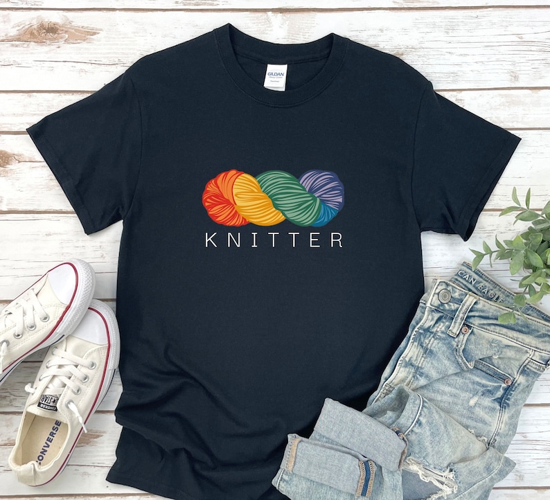 Puede incluir: Camiseta negra con un gr&aacute;fico de ovillo de lana arco&iacute;ris y el texto "KNITTER" impreso en blanco.