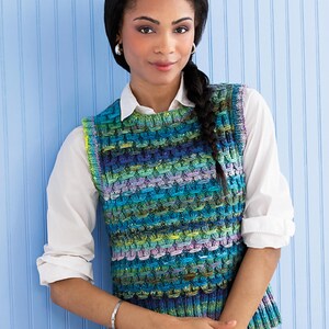 May include: A colorful knitted vest in shades of blue, green, and purple. The vest has a textured pattern and is worn over a white collared shirt. The model is wearing a silver bracelet and ring. The background is a light blue wall.