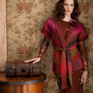 May include: A woman wearing a red, brown and green striped knit cardigan with a brown belt. She is standing in front of floral wallpaper and holding a small wooden box.