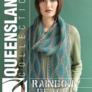 May include: A woman wearing a green shirt is holding a blue and green knitted scarf with a geometric pattern. The scarf is called "Rainbow Beach Kay Scarf" and is part of the "Queensland Collection".