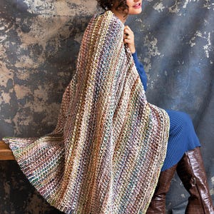 Pode incluir: Um xale multicolorido de tecido aberto drapeado sobre uma pessoa. O xale apresenta uma mistura de cores pastel, incluindo rosa, verde e amarelo. A pessoa está vestindo um vestido azul e botas marrons. O fundo é uma parede cinza texturizada.