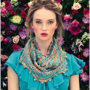 May include: A woman wearing a colorful knitted cowl scarf with blue, yellow, and orange hues. The scarf is draped over a teal blue blouse. The background is a vibrant display of various flowers in shades of pink, red, and white.