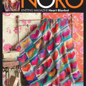 Puede incluir: Una manta de punto de corazón colorida con una variedad de colores, incluyendo rojo, rosa, naranja, amarillo, verde, azul y morado. La manta está drapeada sobre un marco de cama con un papel tapiz de estampado floral.