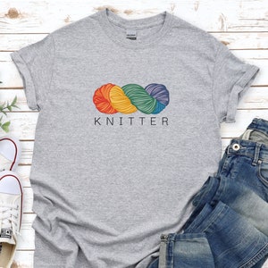 Puede incluir: Camiseta gris con un gr&aacute;fico de ovillo de lana arco&iacute;ris y la palabra "KNITTER" impresa debajo.