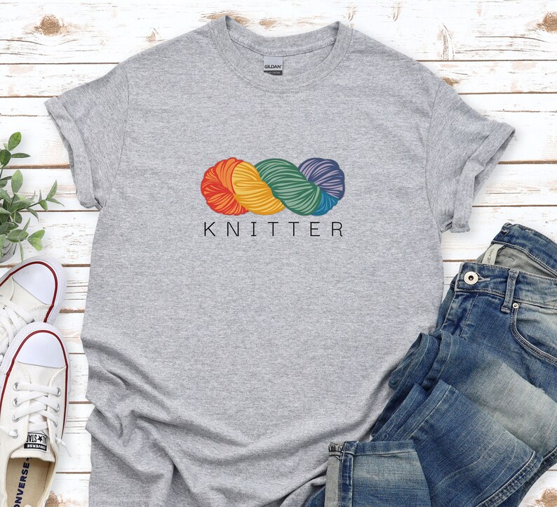 Puede incluir: Camiseta gris con un gr&aacute;fico de ovillo de lana arco&iacute;ris y la palabra "KNITTER" impresa debajo.