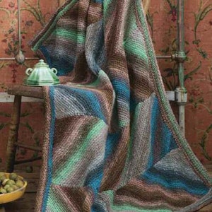 Op de afbeelding: Een veelkleurige gebreide deken, gedrapeerd over een houten stoel. De deken heeft een patchwork-ontwerp met tinten bruin, blauw, groen en grijs. Een kleine groene theepot staat op de stoel. De achtergrond heeft bloemenbehang.