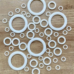 May include: A collection of white, circular O-rings of varying sizes, scattered on a wooden surface. The rings range from small to large, with a smooth, opaque appearance. The arrangement is random, showcasing the different sizes.