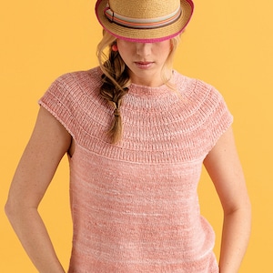 May include: A pink knitted top with short sleeves and a round neckline. The top is made of a soft, lightweight yarn and has a subtle speckled texture. The model is wearing a straw hat with a pink band.