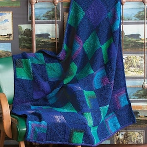 Op de afbeelding: Een gebreide deken met een geometrisch patroon in blauwe, groene en paarse tinten. De deken is over een groene stoel en een metalen bedframe gedrapeerd. De achtergrond toont ingelijste landschapsschilderijen.