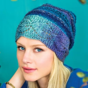 May include: A blue and purple knitted hat with a lacy pattern. The hat is worn by a person with blonde hair.