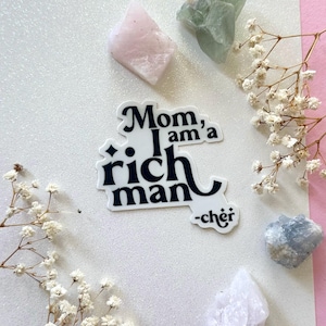 May include: A white sticker with black text that reads "Mom, I am a rich man -chér" on a white background with small white flowers and pink, green, and blue crystals.