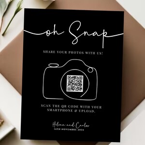 May include: Black and white sign with the text "Oh Snap" and a QR code. The sign says "Share your photos with us!" and "Scan the QR code with your smartphone & upload." The sign also includes the text "Helena and Carlos" and "14th Novembre 2014".