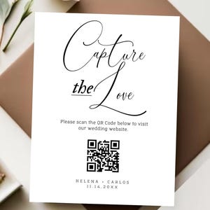 May include: White card with black text that reads "Capture the Love". Below the text is a black and white QR code with the text "Please scan the QR Code below to visit our wedding website." Below the QR code is the text "HELENA CARLOS 11.14.20XX".