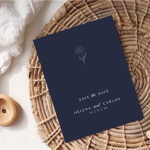 Könnte beinhalten: Eine marineblaue Einladungskarte mit einer floralen Illustration und dem Text "Save the Date". Die Namen "Helena und Carlos" und das Datum "14 | 11 | 35" sind ebenfalls aufgedruckt. Die Karte liegt auf einer geflochtenen Oberfläche.