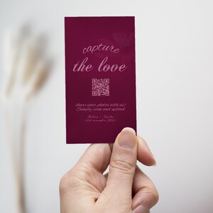 Può includere: Un biglietto bordeaux con il testo "capture the love" e un codice QR. Il biglietto include anche il testo "share your photos with us! Simply scan and upload." Il biglietto è tenuto in mano.