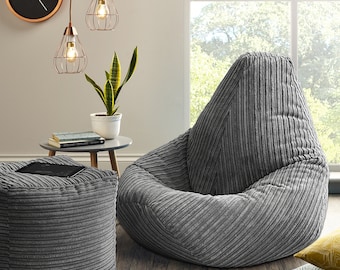 May include: A gray corduroy bean bag chair with a tall back and a wide seat. The chair is sitting on a wooden floor with a gray ottoman and a plant in a white pot in the background.
