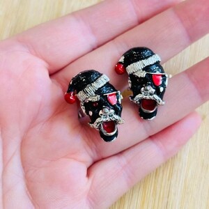May include: A pair of novelty rings featuring black skull faces with Santa hats, red eyes, and open mouths. The rings have silver-colored accents and are set against a light background.