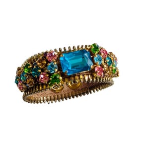 Könnte beinhalten: Ein goldfarbenes Armband mit einem großen, rechteckigen, türkisfarbenen Edelstein in der Mitte. Das Armband ist mit einer Reihe von bunten, kleineren Edelsteinen in Rosa, Grün und Blau verziert. Das Design beinhaltet eine reißverschlussartige Textur.