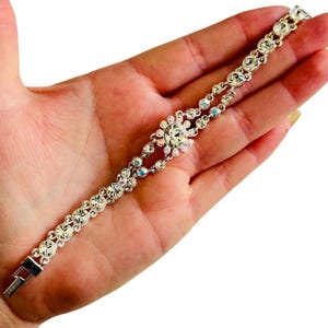 Puede incluir: Una pulsera plateada con brillantes redondos y transparentes y un diseño floral central. La pulsera presenta un diseño de doble hilera y un cierre seguro. Los brillantes brillan. Una delicada pieza de joyería.