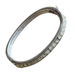 Puede incluir: Un brazalete articulado de color plateado. El brazalete presenta una fila de cristales cuadrados y transparentes a lo largo del borde superior. El cierre es visible en la parte superior. El brazalete está sobre un fondo blanco.