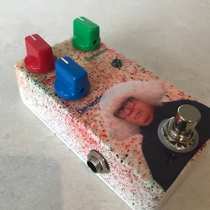 May include: A white guitar effects pedal with a red, green, and blue knob. The pedal has a splatter paint design with red and green paint splatters. The pedal has a picture of a man with a white hat and glasses on it. The pedal has a black and silver footswitch.