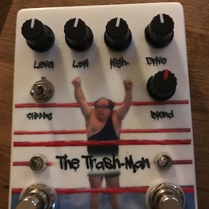 Puede incluir: Pedal de efectos de guitarra blanco con temática de lucha libre. El pedal tiene cuatro perillas etiquetadas como "Level", "Low", "High" y "Drive". Hay un interruptor de palanca etiquetado como "Clipping" y una perilla etiquetada como "Blend". El pedal presenta un gráfico de un luchador en un ring con el texto "The Trash-Man".