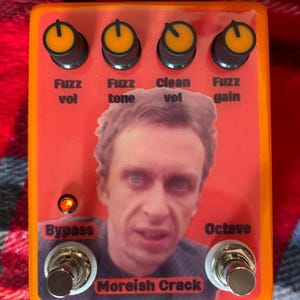 Może przedstawiać: Pomarańczowy pedał efektów gitarowych z czterema pokrętłami oznaczonymi jako "Fuzz vol", "Fuzz tone", "Clean vol" i "Fuzz gain". Pedał ma zdjęcie twarzy mężczyzny i napis "Moreish Crack". Posiada również przełączniki "Bypass" i "Octave".