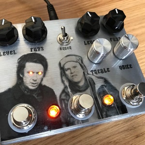 Puede incluir: Un pedal de efectos de guitarra plateado con botones negros y un gráfico de dos hombres. El pedal tiene las palabras "Level", "Fuzz", "Order", "Treble" y "Voice" impresas en él. El pedal tiene dos luces LED naranjas.
