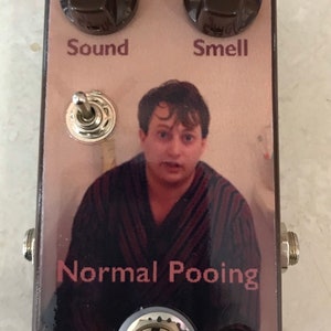 Puede incluir: Un pedal de efectos de guitarra personalizado con una cara marrón que presenta a un hombre con una bata. El pedal tiene dos perillas marrones con las etiquetas "Sound" y "Smell", un interruptor de palanca y el texto "Normal Pooping".