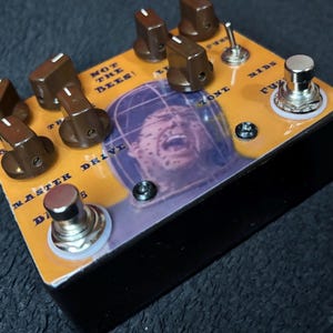 Puede incluir: Un pedal de efectos de guitarra rectangular con perillas marrones y un interruptor de pie plateado. El pedal presenta el texto "NOT THE BEES!" y un gráfico de la cara de una persona cubierta de abejas. El pedal también tiene el texto "MASTER DRIVE".