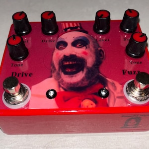 May include: A red guitar effects pedal with a graphic of a clown's face. The pedal has black knobs with red tops, labeled "Tone", "Drive", "Level", and "Fuzz". It also has a silver foot switch.