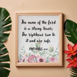 Può includere: Stampa incorniciata con il testo "The name of the Lord is a strong tower; The righteous run to it and are safe. PROVERBS 18:10." La stampa presenta un'arte floreale ad acquerello in rosa e bianco.