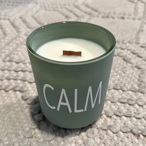 Könnte beinhalten: Ein grünes Glas-Kerzengefäß mit einem weißen Etikett, auf dem "CALM" steht. Die Kerze ist angezündet und hat einen Holzdocht.