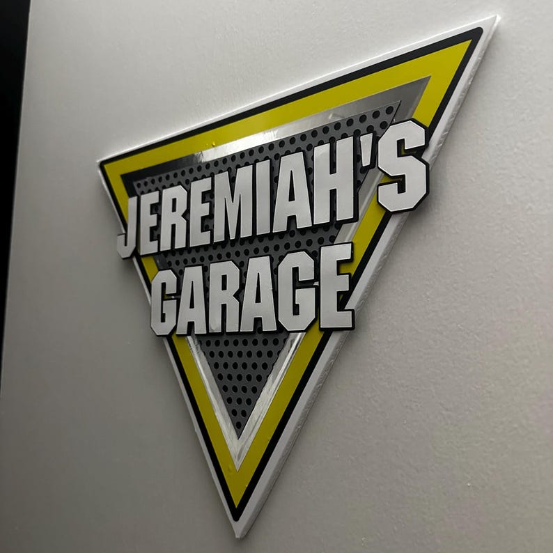 Puede incluir: Un letrero triangular con el texto "JEREMIAH'S GARAGE" en blanco, delineado en negro, sobre un fondo gris. El letrero tiene un borde amarillo y est&aacute; montado en una pared blanca.