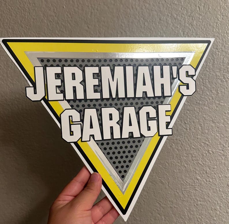 Puede incluir: Un letrero triangular con el texto "JEREMIAH'S GARAGE" en letras blancas en negrita. El letrero tiene un borde amarillo y negro, con un fondo gris punteado. El letrero est&aacute; sostenido por una mano.
