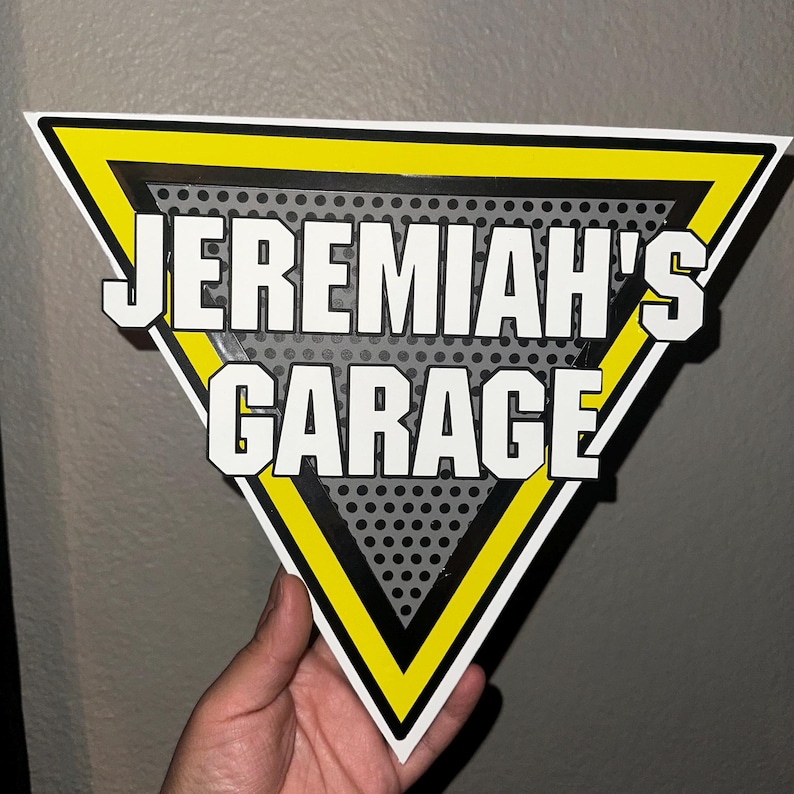 Puede incluir: Un letrero triangular con el texto "JEREMIAH'S GARAGE" en letras blancas. El letrero tiene un borde amarillo y un fondo gris punteado. El letrero est&aacute; sostenido por una mano.