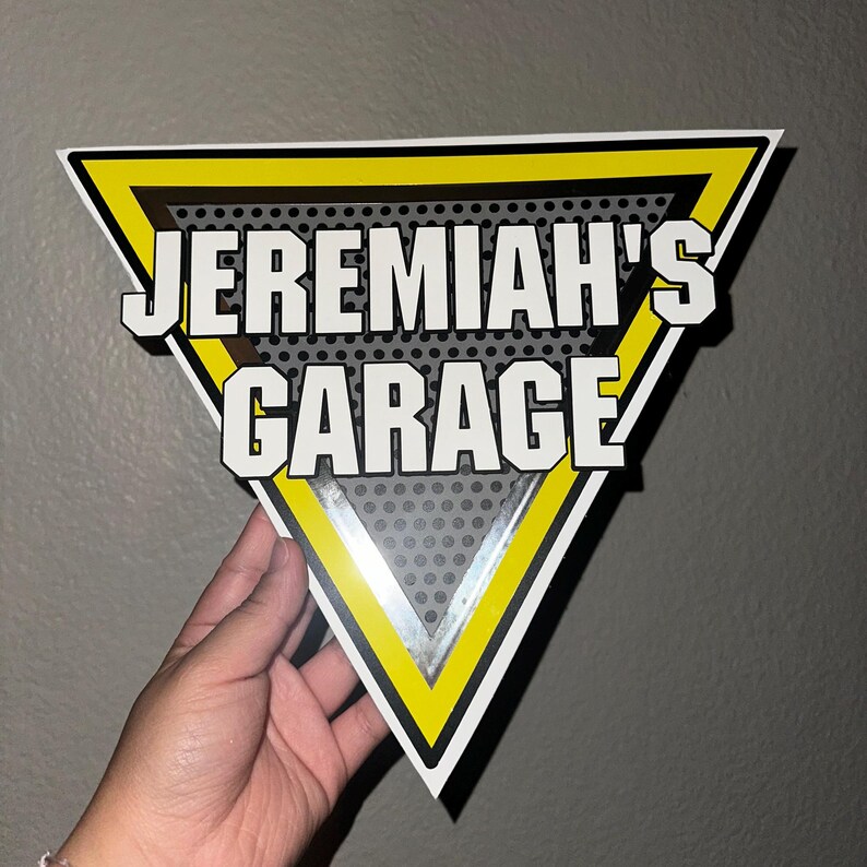 Puede incluir: Un letrero triangular con el texto "JEREMIAH'S GARAGE" en blanco, delineado en negro. El letrero tiene un borde amarillo y negro y un fondo gris perforado. El letrero est&aacute; sostenido por una mano.