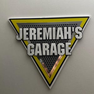 Puede incluir: Un letrero triangular con el texto "JEREMIAH'S GARAGE" en letras blancas en negrita con contornos negros. El letrero tiene un borde amarillo, un interior plateado y un fondo gris perforado.