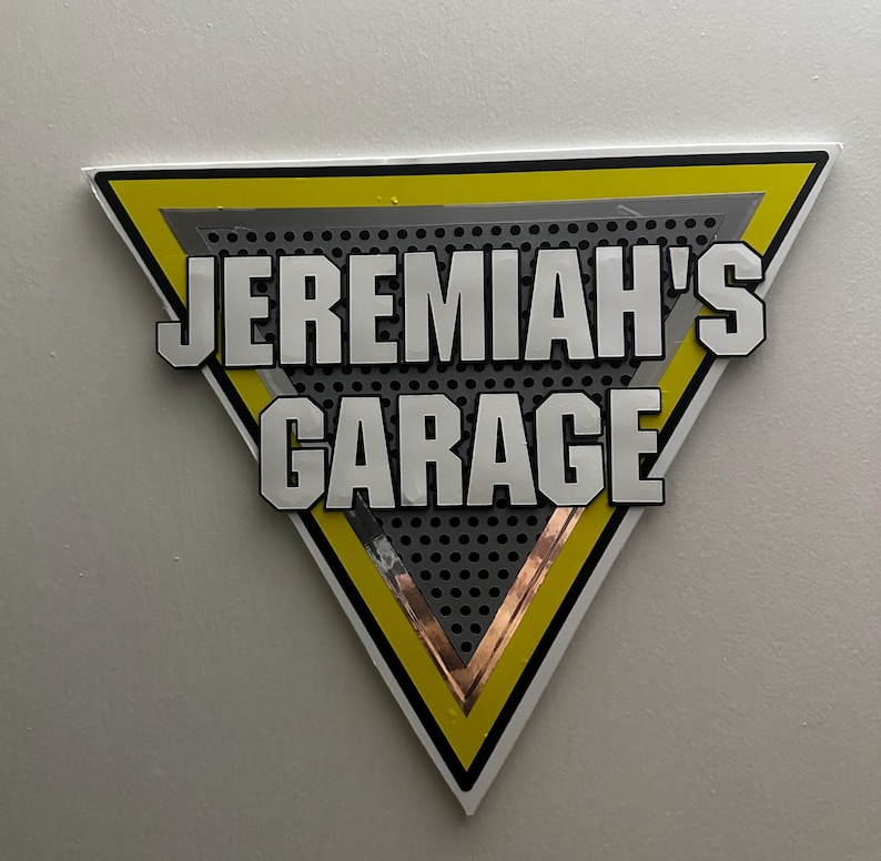 Puede incluir: Un letrero triangular con el texto "JEREMIAH'S GARAGE" en letras blancas en negrita con contornos negros. El letrero tiene un borde amarillo, un interior plateado y un fondo gris perforado.
