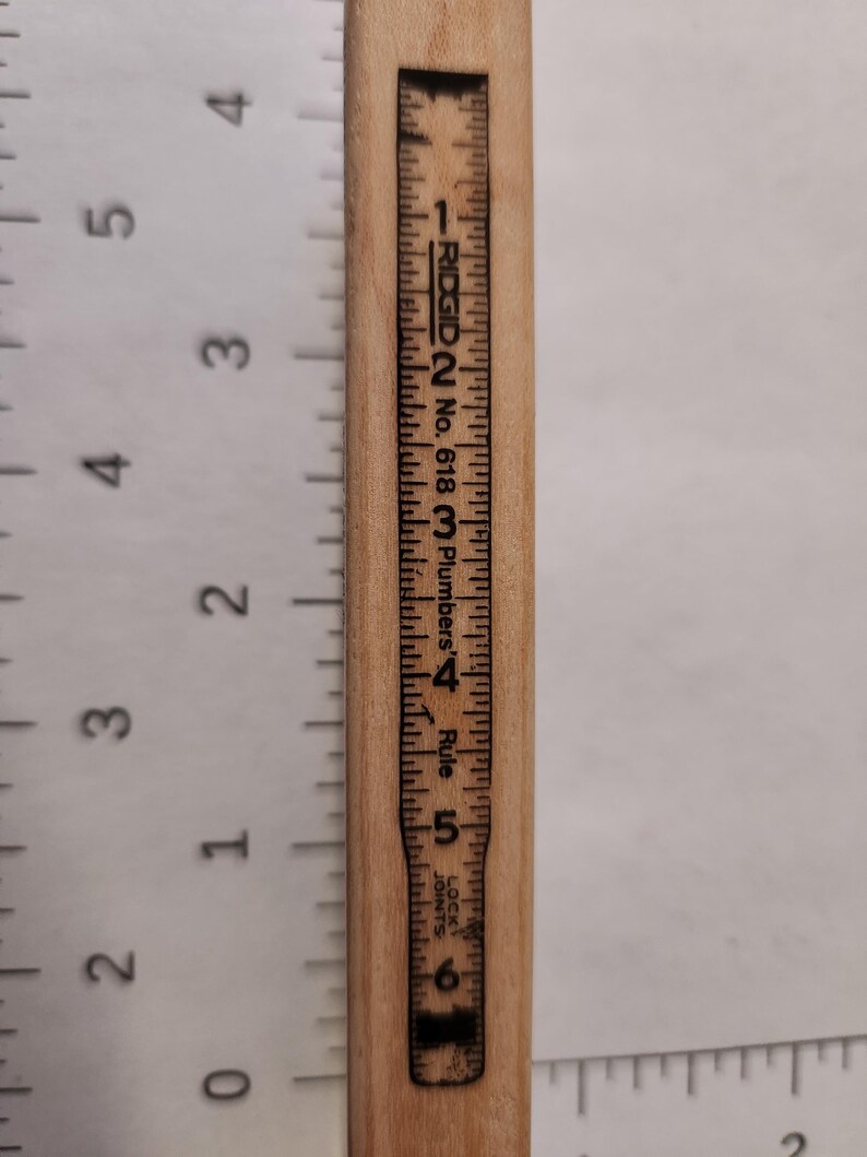 May include: A wooden ruler with black markings and the text "RIDGID No. 618 Plumbers' Rule." The ruler measures up to 6 inches. The numbers and markings are printed in black ink on the light brown wood.
