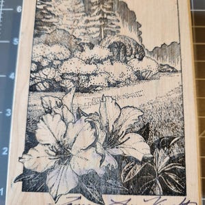 May include: A wooden stamp featuring a detailed black and white illustration of a landscape with flowers in the foreground. The scene includes a field, trees, and a mountain range. The stamp is signed "Tamara Lee Knight".