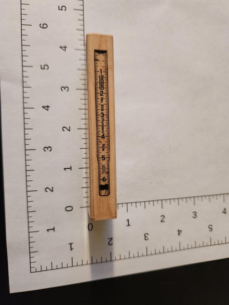 May include: A small, rectangular wooden ruler with black markings and numbers. The ruler is marked with inches and has the text "RIDGID No. 618 Plumbers Rule" printed on it. The ruler is approximately 6 inches long.