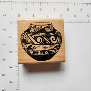 May include: A wooden stamp featuring a black and white illustration of a vase with intricate tribal designs. The stamp is rectangular and has a detailed image of the vase, perfect for crafting or art projects.
