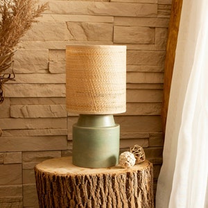 Pode incluir: Um abajur de tecido trançado com uma base verde fica em cima de uma mesa de cabeceira de tronco de árvore ao lado de uma cama com lençóis brancos e um dossel branco transparente.