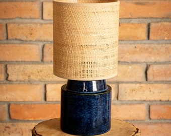 May include: A blue ceramic table lamp with a woven natural fiber shade. The lamp is sitting on a wooden stump.